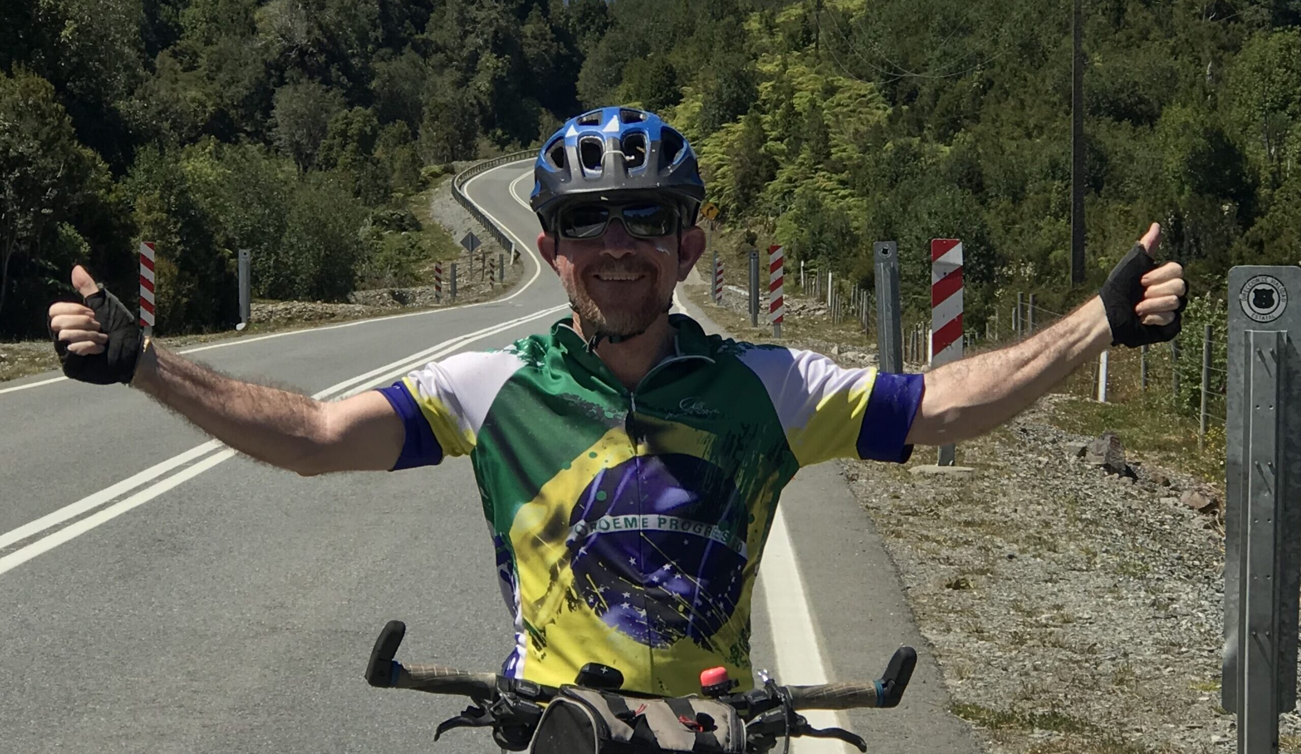 Ciclista sorridente com uniforme do Brasil fazendo sinal de positivo com os dois braços em estrada sinuosa cercada por vegetação nativa, durante uma viagem de cicloturismo.