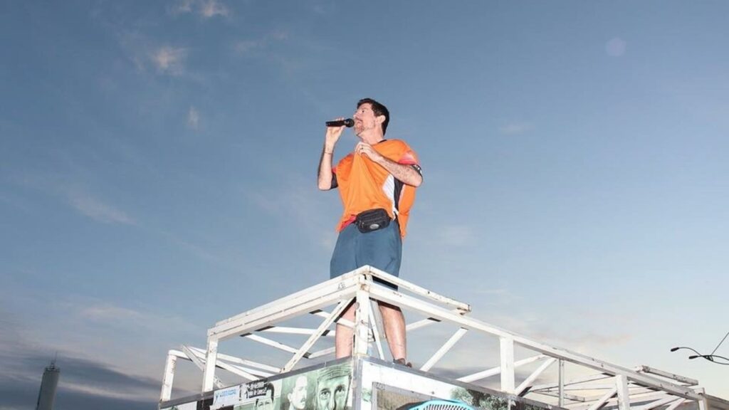 Cicloativista Aparecido Inácio de camiseta laranja fala ao microfone em cima de uma estrutura metálica durante evento ao ar livre, com o céu limpo ao fundo. A cena transmite engajamento e liderança em prol da mobilidade urbana por bicicleta.