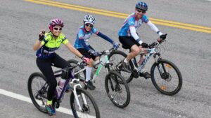 Três ciclistas sorrindo pedalam juntos em uma estrada, representando a prática do ciclismo em família como forma de promover saúde e bem-estar.