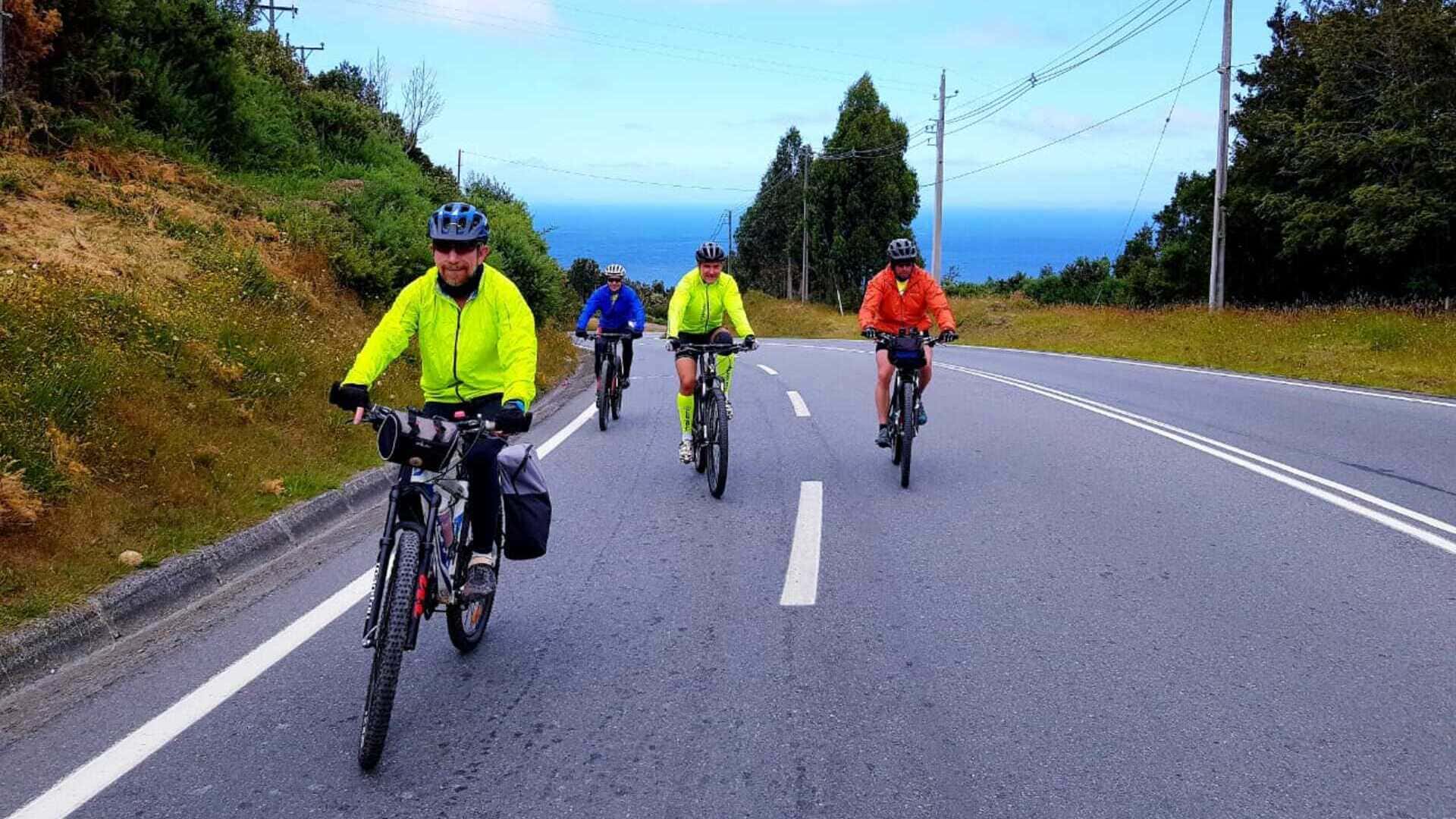 Grupo de cicloturistas pedalando em estrada asfaltada com vista para o mar, usando roupas coloridas e equipamentos de cicloviagem, representando aventura, turismo sustentável e conexão com a natureza.