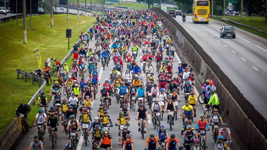 Imagem do Pedal Anchieta 2019 com milhares de ciclistas ocupando a rodovia em São Paulo durante o maior evento de ciclismo urbano do Brasil. A multidão de ciclistas com roupas coloridas e capacetes representa o fortalecimento do cicloativismo e o incentivo ao uso da bicicleta como meio de transporte sustentável.