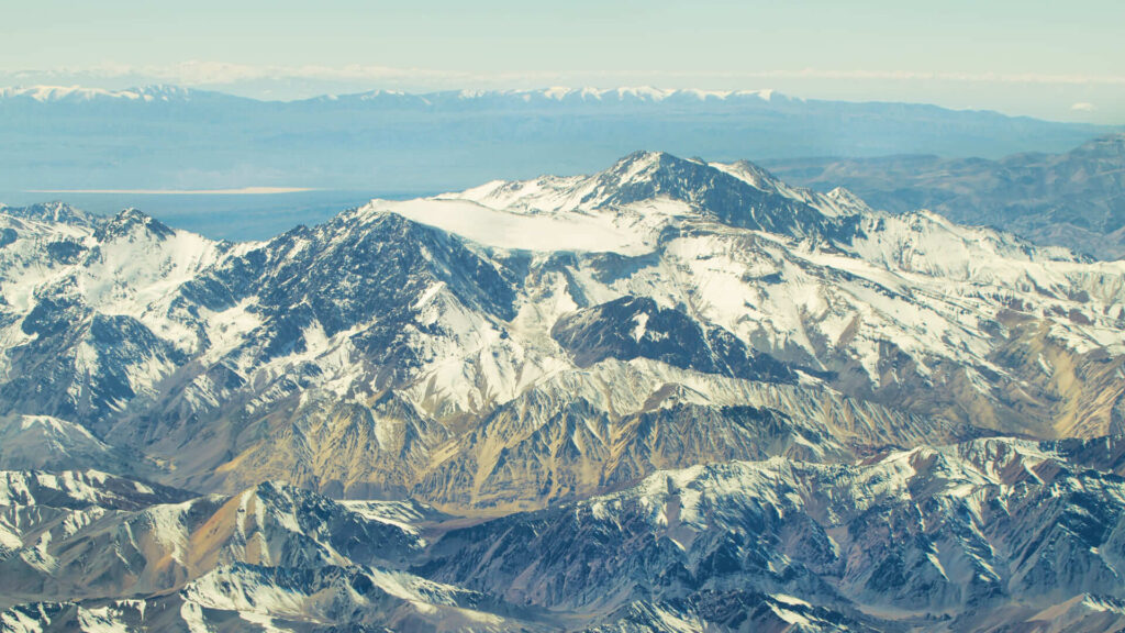 Vista aérea da Cordilheira dos Andes, com montanhas imponentes cobertas de neve e vales áridos em contraste, revelando a grandiosidade e diversidade da paisagem andina.