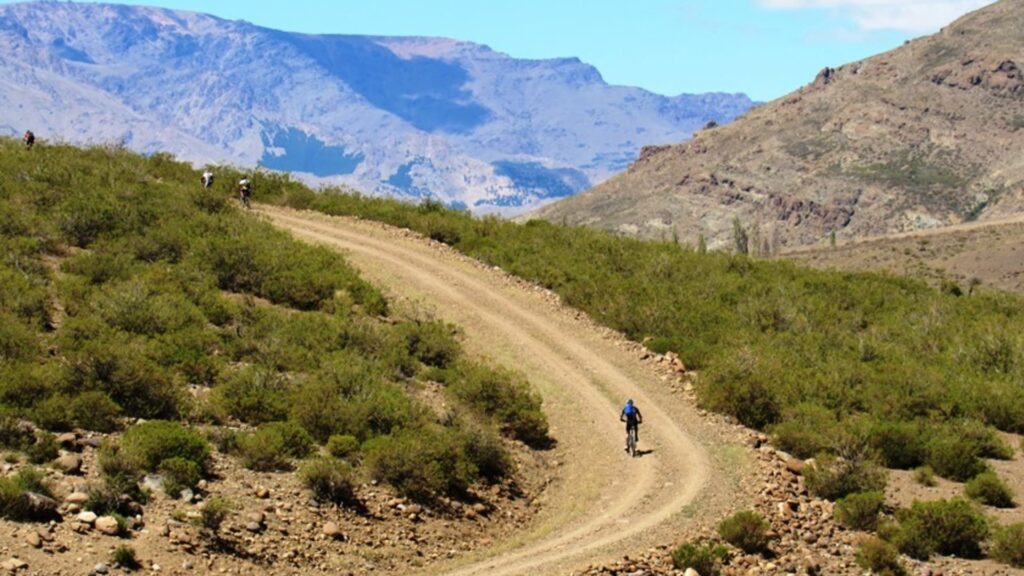 Ciclista pedalando em uma estrada de terra sinuosa na Cordilheira dos Andes, rodeado por vegetação baixa e montanhas rochosas ao fundo, sob céu azul claro.