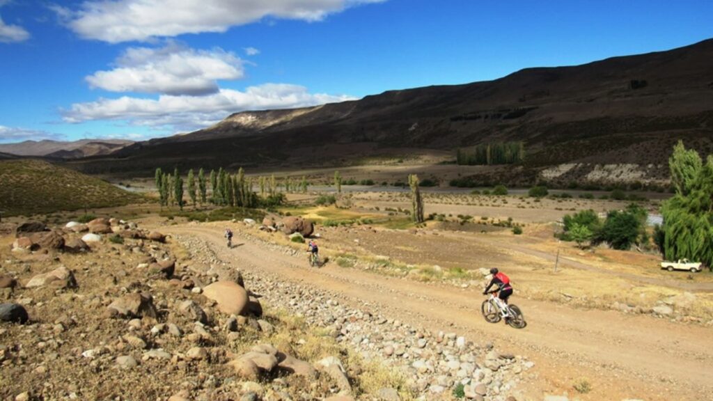 Ciclistas pedalando por uma estrada de terra na Cordilheira dos Andes, cercados por um cenário árido com rochas, vegetação esparsa e montanhas ao fundo sob um céu azul com nuvens.