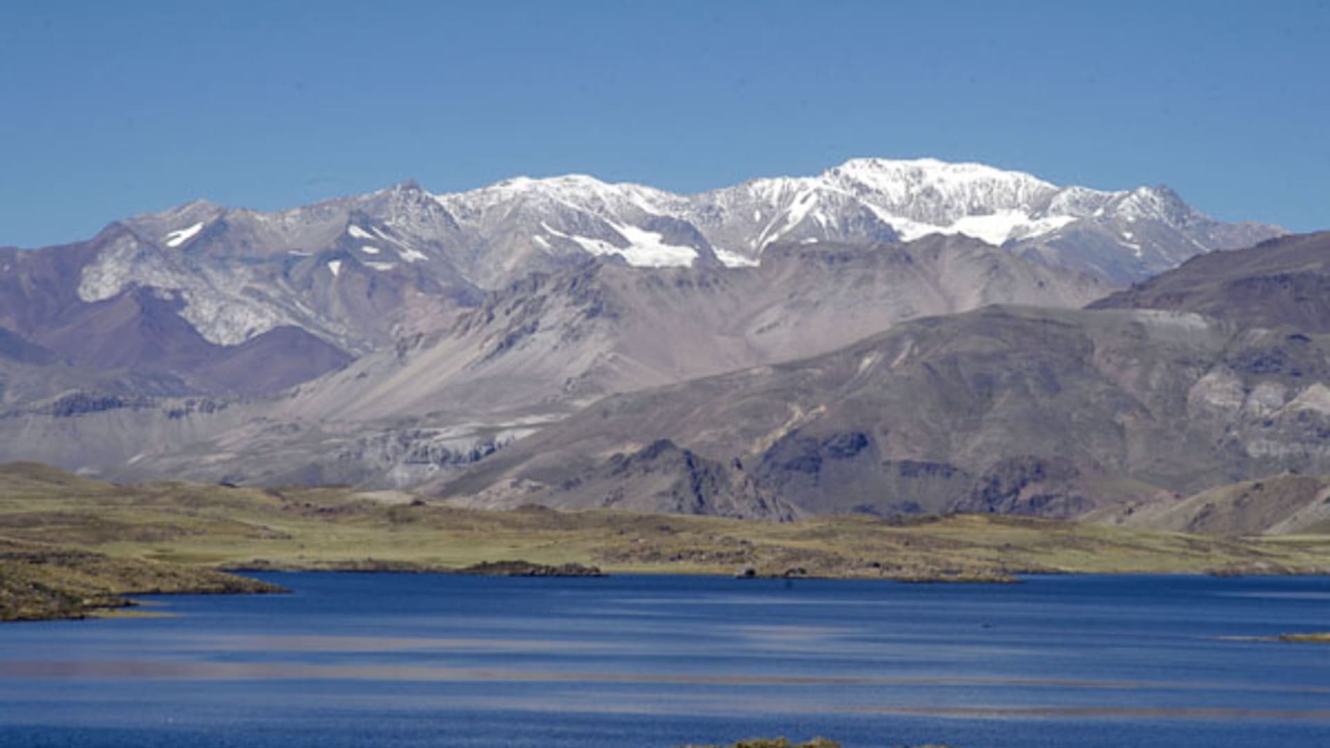 Paisagem da Cordilheira dos Andes com montanhas imponentes, algumas cobertas de neve no topo, em contraste com o lago azul em primeiro plano e vales verdes ao redor.