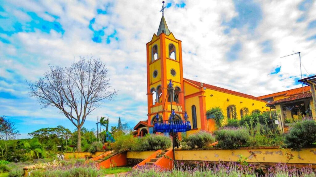 Igreja amarela com detalhes laranja rodeada por jardim com lavandas e árvores, sob céu azul com nuvens, em cenário tranquilo e pitoresco.
