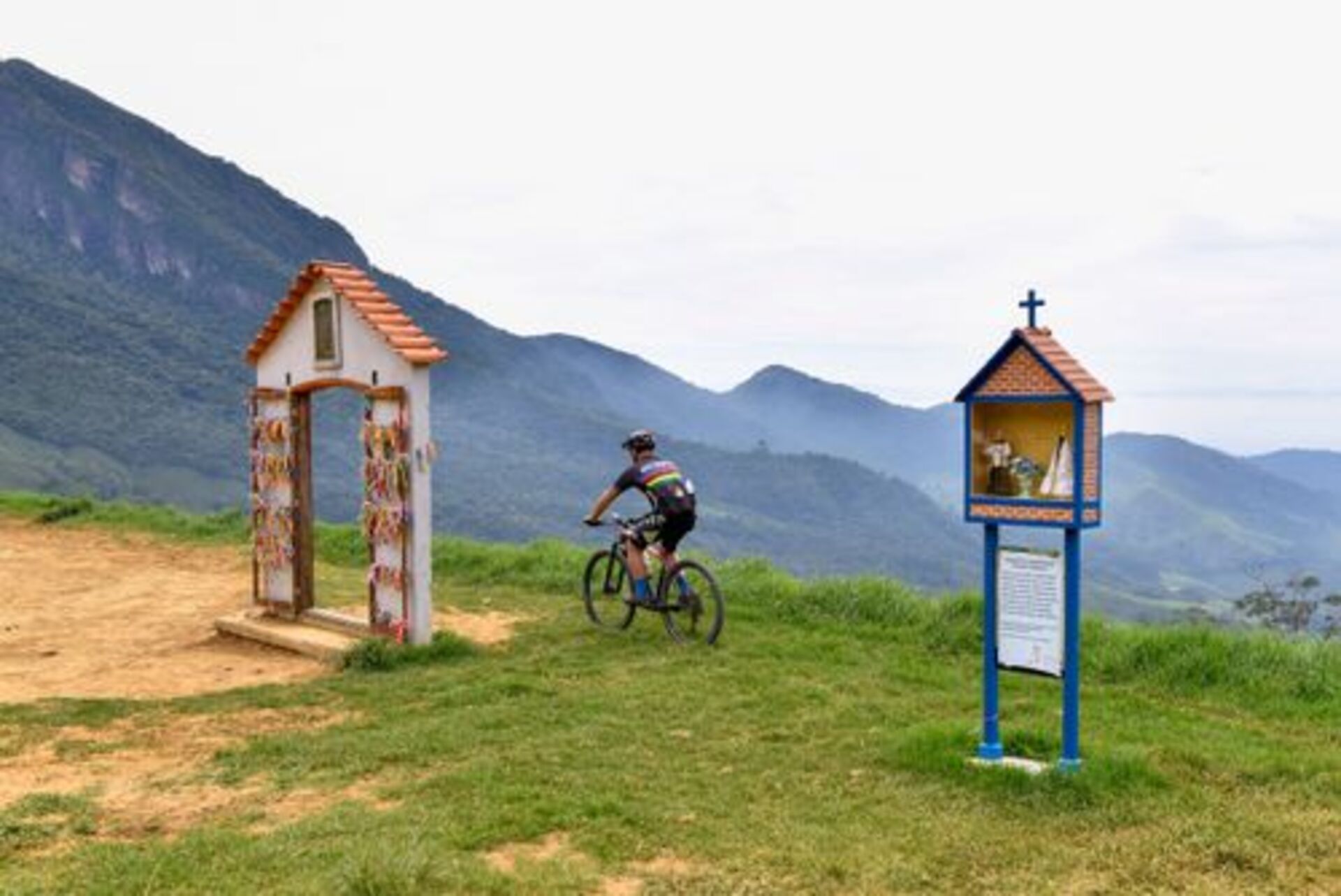 Ciclista pedalando em trilha de montanha, passando por portal decorado com fitas coloridas e pequeno oratório religioso, com vista panorâmica para serras verdes ao fundo.