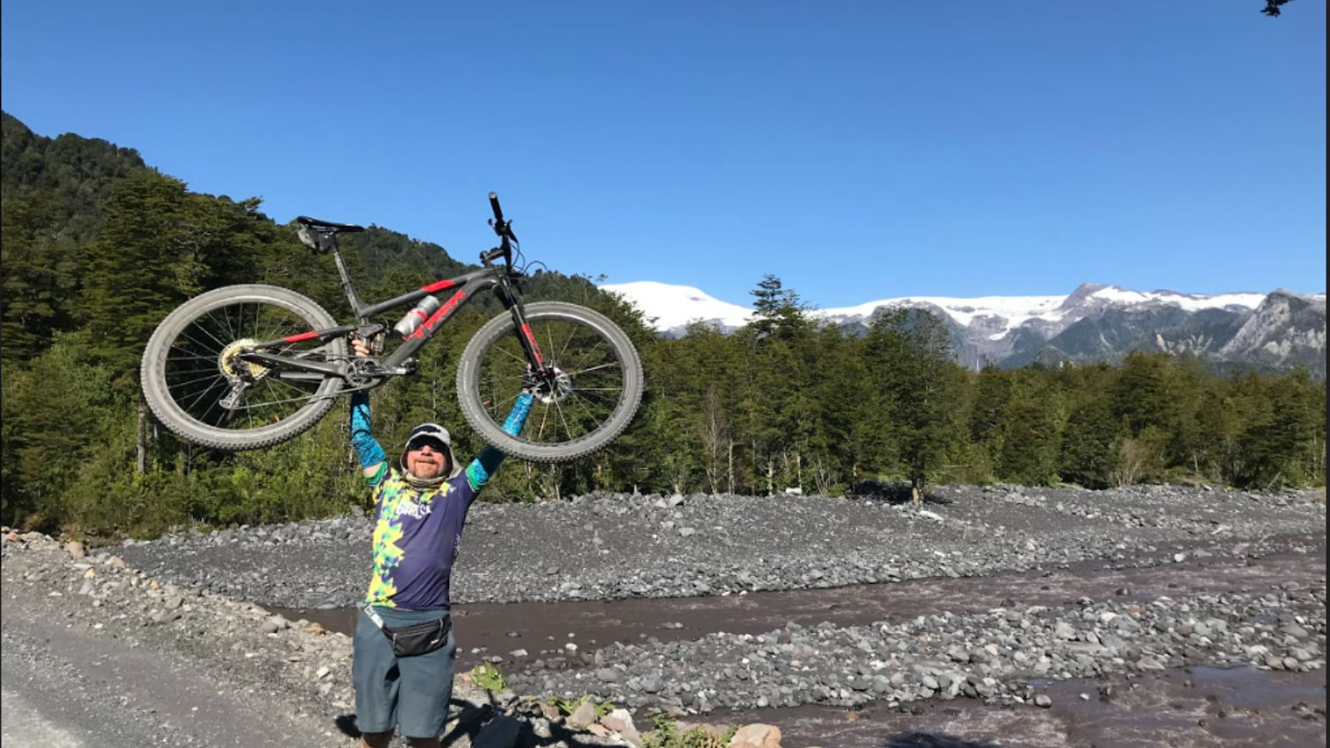 Homem levantando bicicleta de montanha acima da cabeça em um rio, com floresta e montanhas cobertas de neve ao fundo, durante expedição de cicloturismo na Cordilheira dos Andes, Patagônia Argentina.
