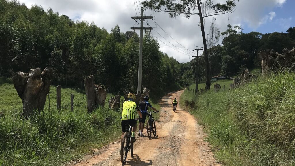Grupo de ciclistas pedalando em estrada de terra cercada por vegetação e árvores, em um passeio de cicloturismo rural.